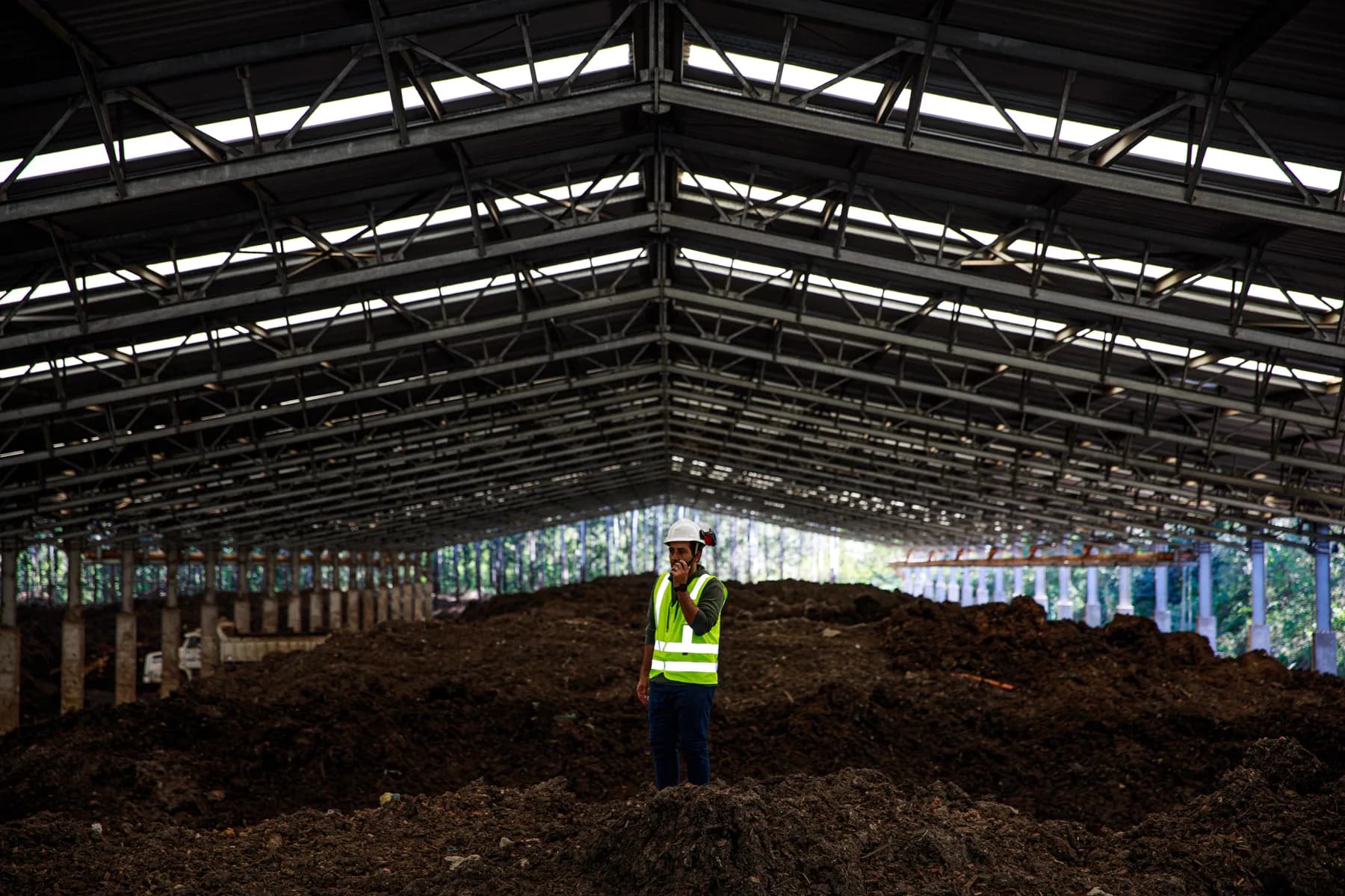 A worker in a hi-vis vest and white helmet standing among compost windrows inside the covered shed at Tera Ambiental