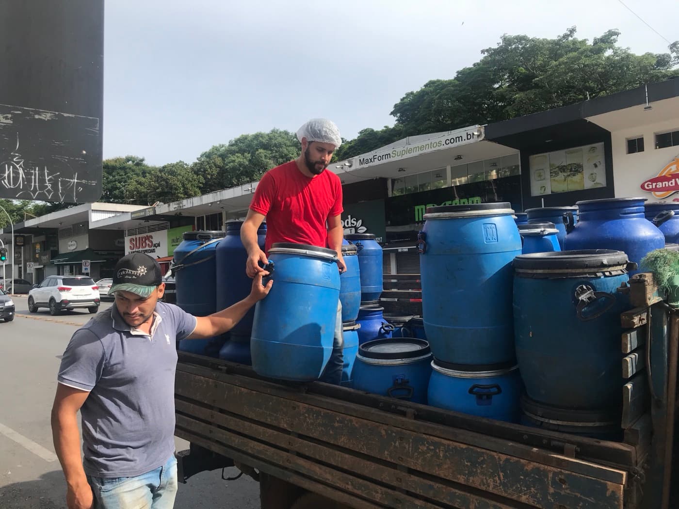 Equipe de coleta carregando bombonas azuis de resíduos alimentares em um caminhão diante de restaurantes de Brasília
