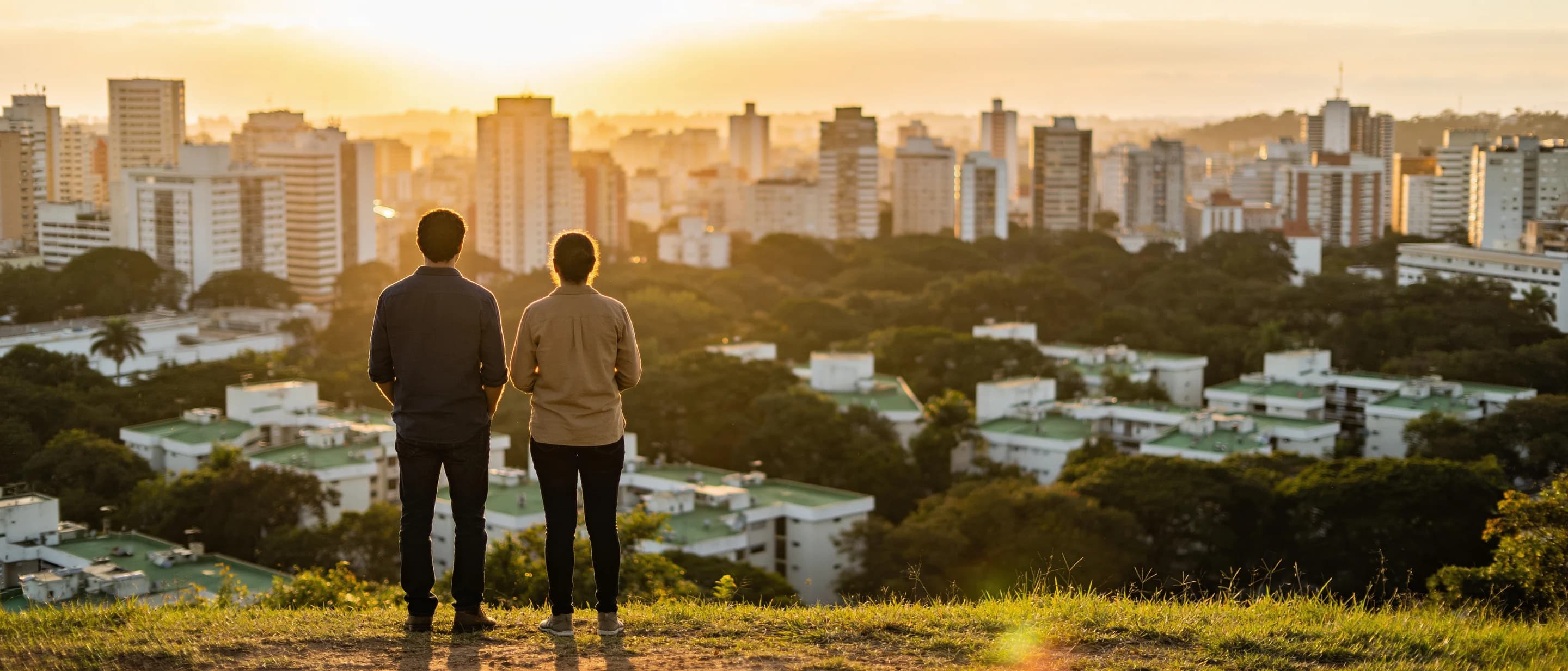 Two people stand on a grassy overlook at sunset, looking out over a Brazilian city where dense tree canopy grows between the buildings.